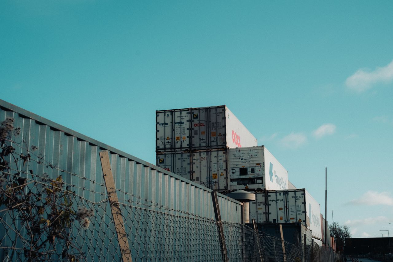 The Art of Drawing Readers In: Your attractive post title goes here A view of stacked shipping containers beside a fence under a clear blue sky, captured outdoors.